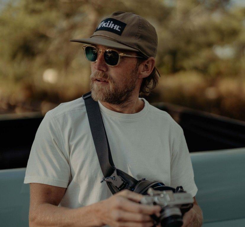 A man holding a camera wearing a KUHL Renegade Camp Hat and sunglasses